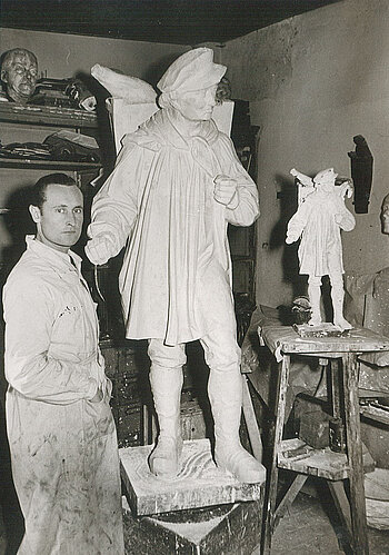 Von links nach rechts: Der Bildhauer, ein junger Mann, in weißem Kittel in seiner Werkstatt. Das große Gipsmodell des Kiepenkerldenkmals, so groß wie der Bildhauer aber auf einem Sockel. Das kleine Modell des Kiepenkerldenkmals auf einem hohen Tisch auf augenhöhe. 