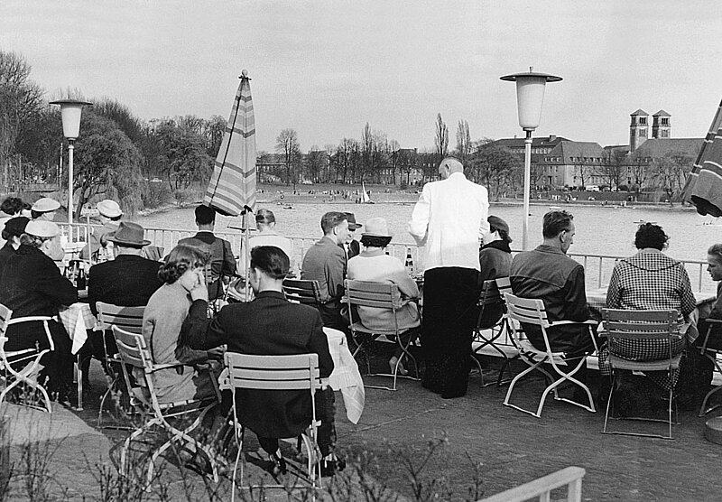 Schwarzweißfoto: Menschen sitzen an Tischen eines Cafés am Aasee in Münster.