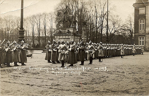 Schwarzweißfoto auf einer Postkarte. Soldaten in Paradeuniform aufgestellt vor einem Reiterstandbild. Einige spielen Instrumente. 