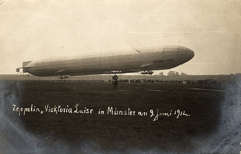 Schwarzweißbild: Luftschiff Viktoria Luise über einer Wiese, davor ein Reiter.. Text auf dem Bild: Zeppelin "Vicktoria Luise in Münster am 9. Juni 1912